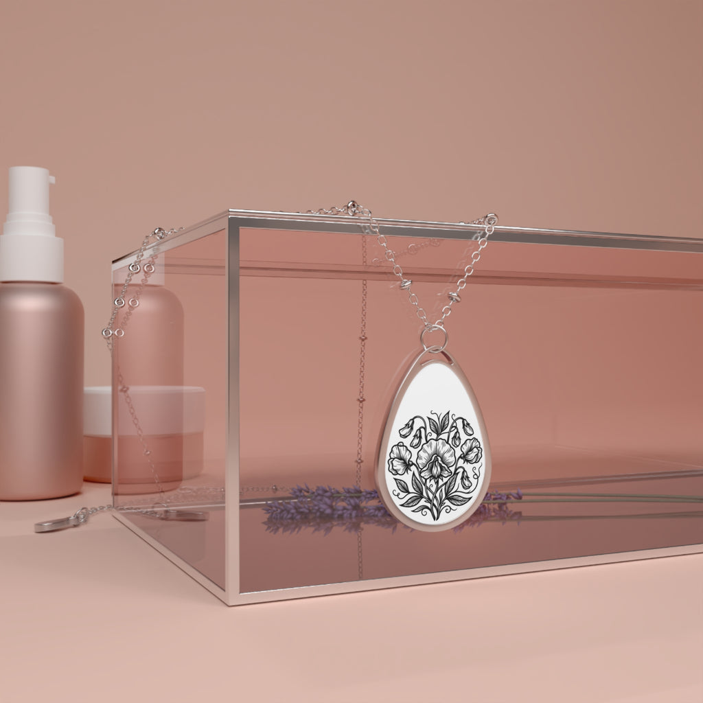 Teardrop-shaped pendant with floral design on a chain, placed inside a clear box with a pink background.