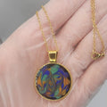 Gold necklace with a colorful pendant held in a hand against a neutral background
