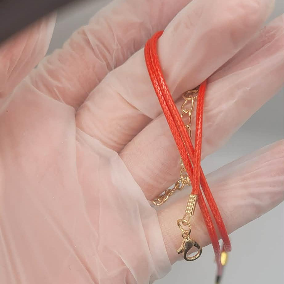 Hand wearing a clear glove holding a red string with a gold charm against a neutral background