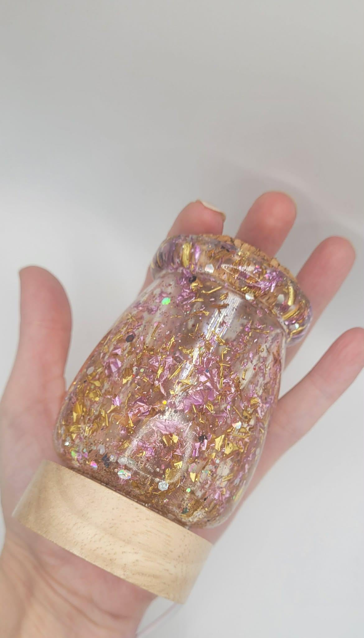Hand holding a clear bottle filled with glittery pink and gold contents against a white background